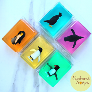 Five colorful penguin soaps in clear plastic clamshell packaging, arranged on a white marble surface. The soaps are in vibrant shades of orange, yellow, turquoise, pink, and green, with black, white, and yellow plastic toy 
penguin figures  inside.