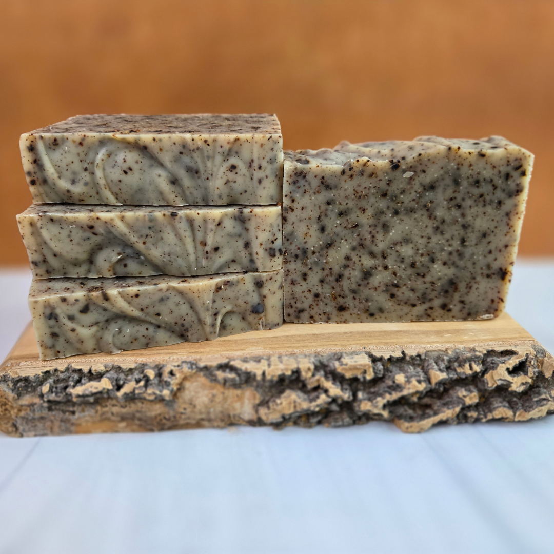 Three stacked bars of soap along side one bar standing and facing forward. The soaps are tan in color with brown speckles. The soaps are on a slab of wood with bark showing and the background is gold.