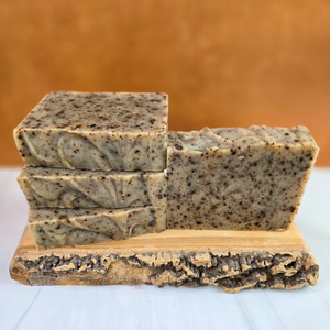 Three rectangular bars of handmade soap with a speckled gray and beige texture, displayed on a rustic bark-edged wooden block against a warm brown background.